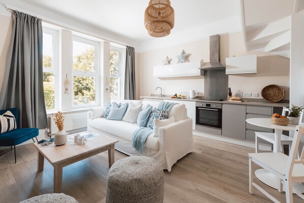 Ein modernes Wohnzimmer mit einer Couch, einem Couchtisch und einem Hocker. Im Hintergrund ist eine Küche mit Herd, Backofen und Schränken zu sehen.