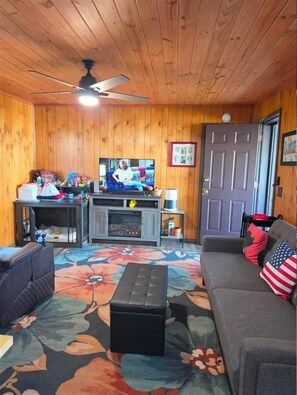 Ein gemütliches Wohnzimmer mit Fernseher, Sofa, Hocker und Deckenventilator.