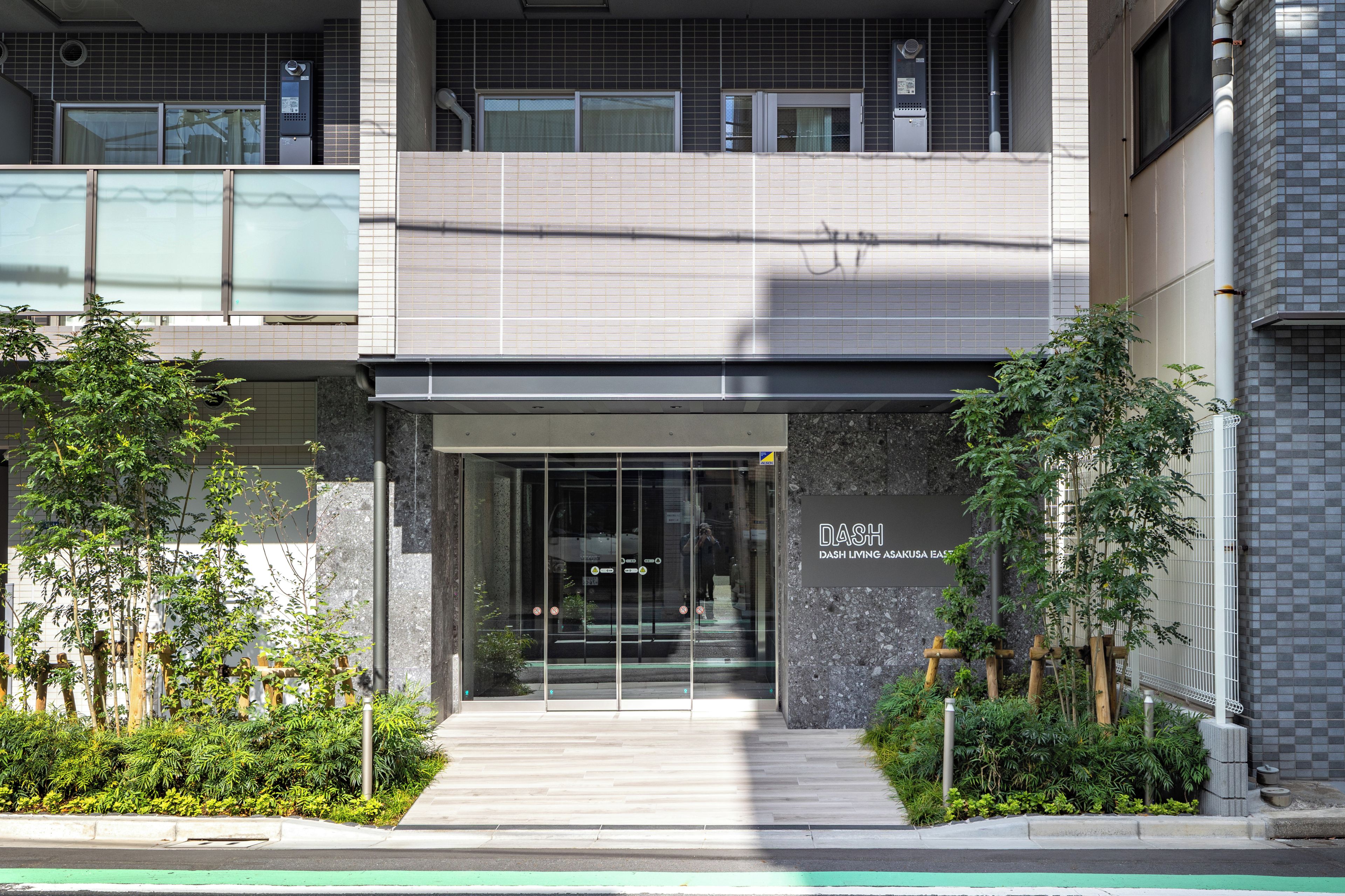 A modern building entrance with a glass door, a sign reading 'DASH Living Asakusa East,' and a small garden area with plants.