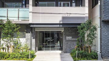 A modern building entrance with a glass door, a sign reading 'DASH Living Asakusa East,' and a small garden area with plants.