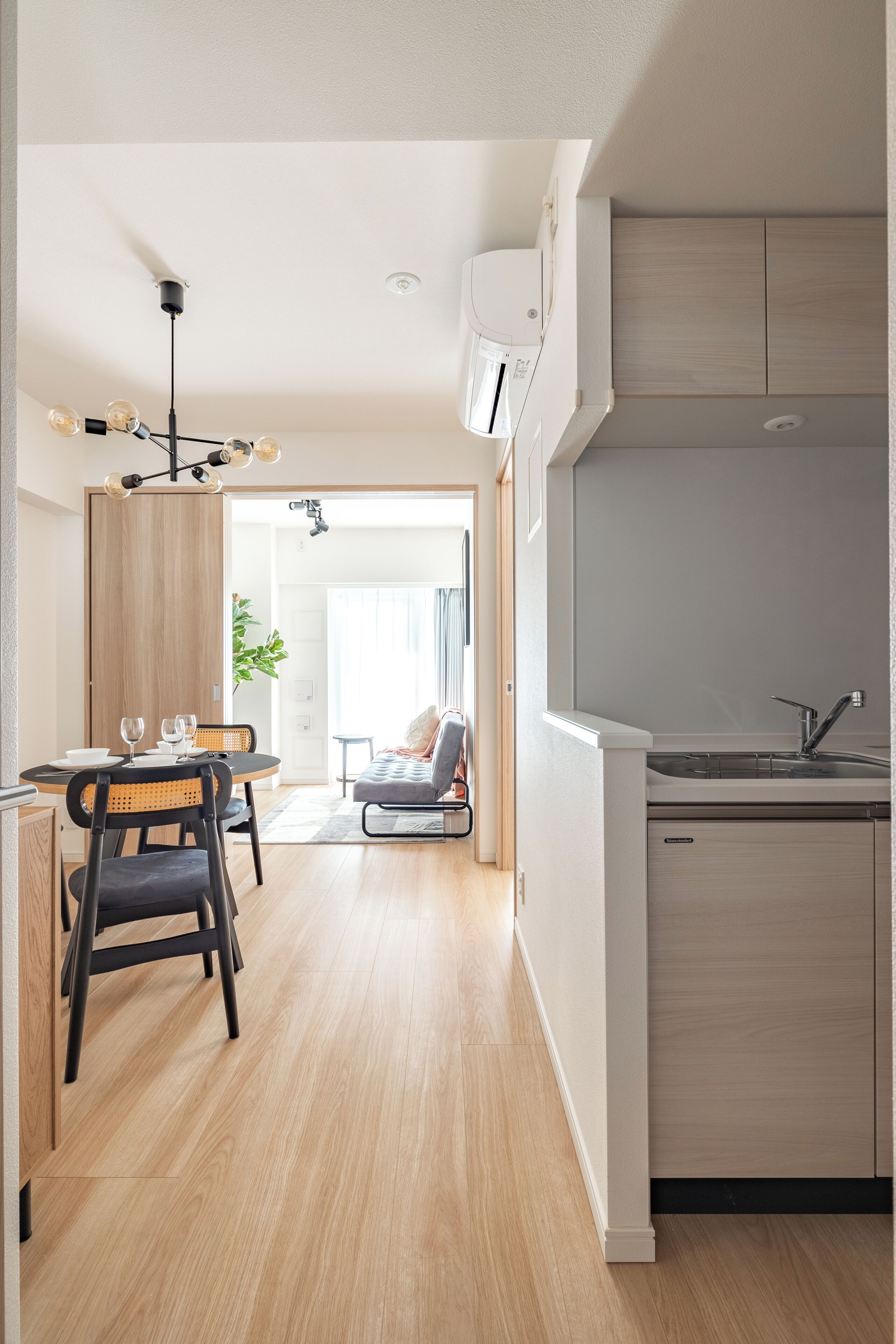 A modern kitchen with a built-in dishwasher, a dining area with a wooden table and chairs, and a living space with a sofa and a plant.