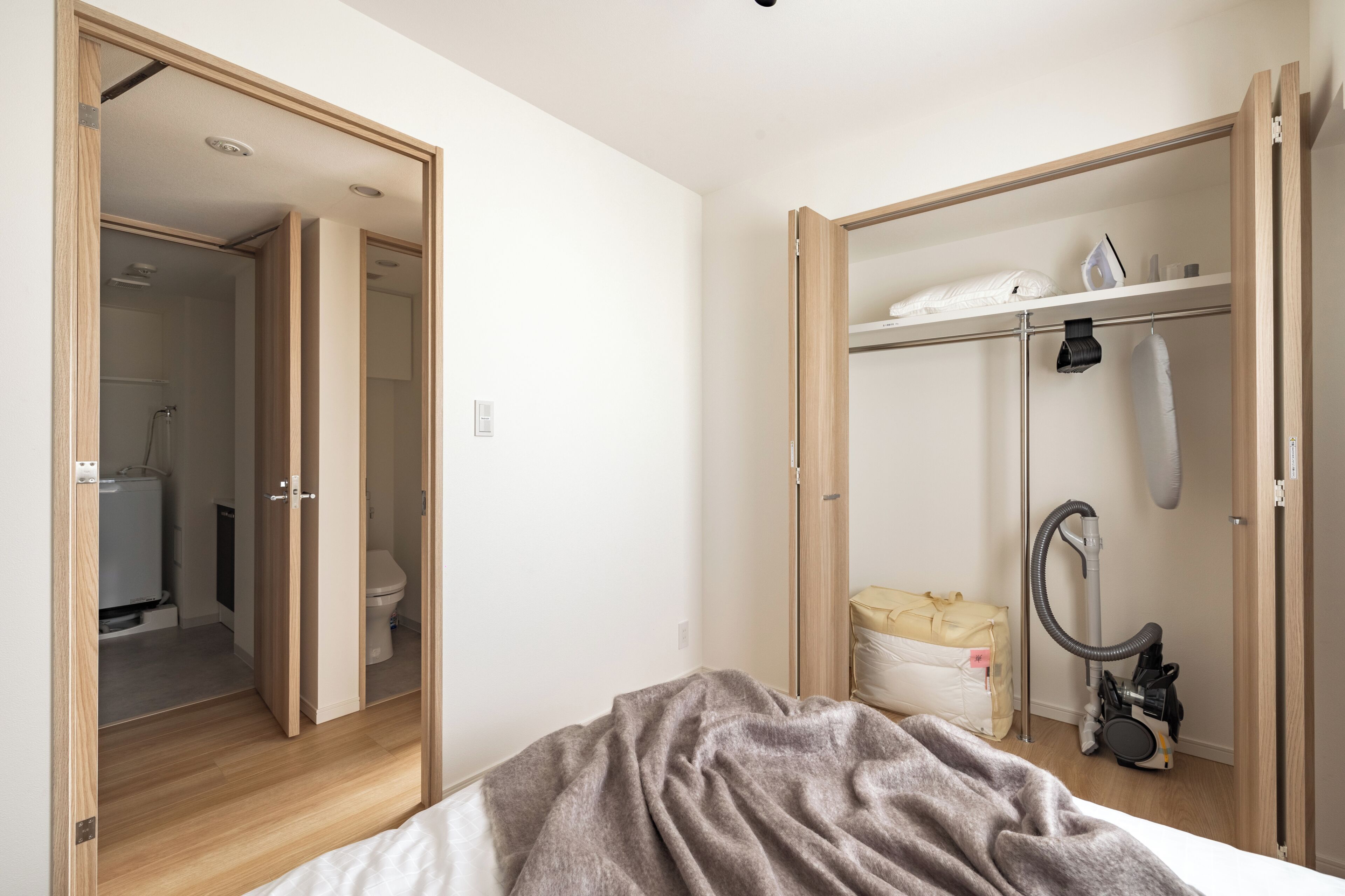 A bedroom with a bed, a closet with a hanging iron, and a vacuum cleaner.