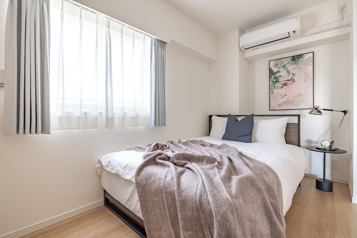 A bedroom with a bed, a bedside table with a lamp, a wall-mounted air conditioner, and a window with curtains.
