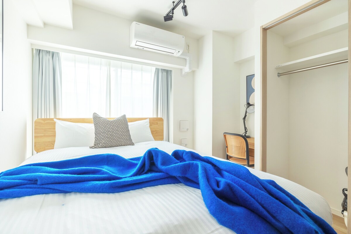 A hotel room with a bed, a window with curtains, an air conditioning unit, a desk with a chair, and a door.
