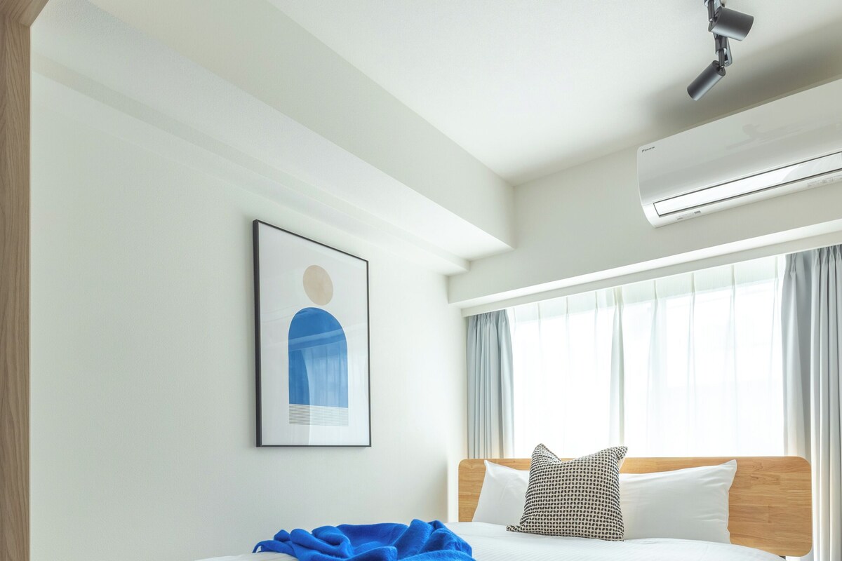 A hotel room with a bed, a headboard, a window with curtains, an air conditioning unit, and a framed picture on the wall.