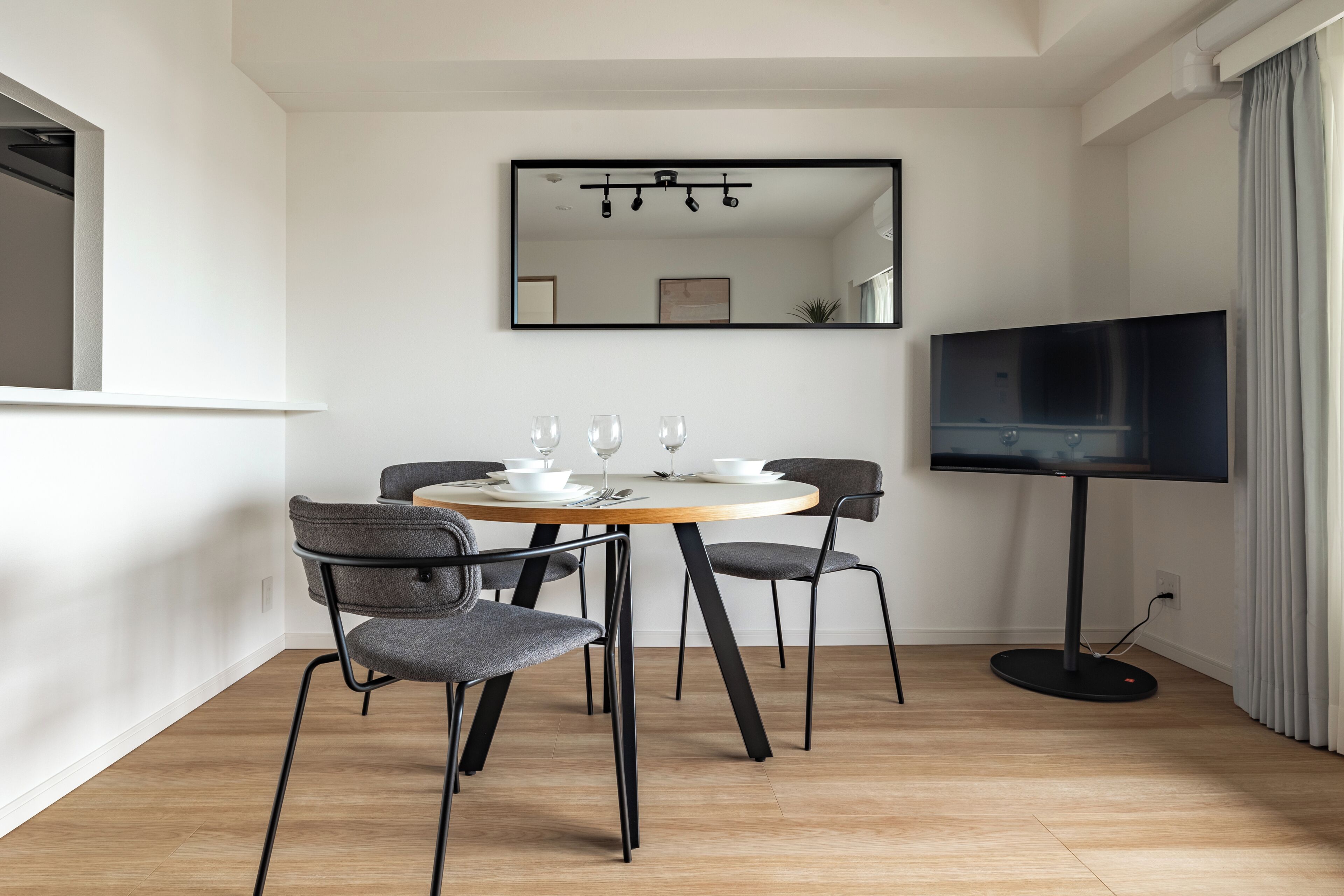 A modern dining area with a round table, four chairs, a flat-screen TV, and a mirror on the wall.