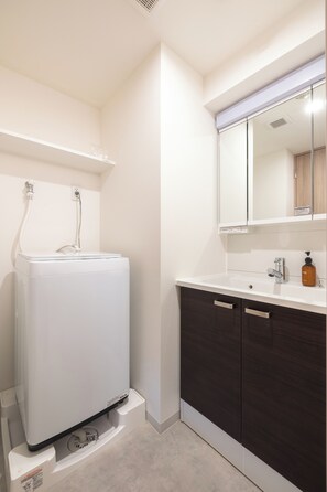 A bathroom with a white washing machine, dark wooden cabinets, a sink with a mirror, and a bottle of soap. - Dash Living Asakusa East (Higashikomagata, Sumida City)