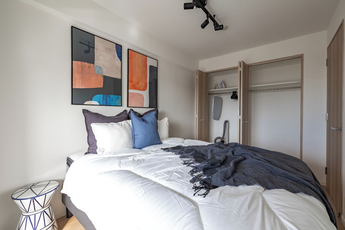A bedroom with a bed, pillows, a blanket, a wardrobe, and abstract art on the wall.