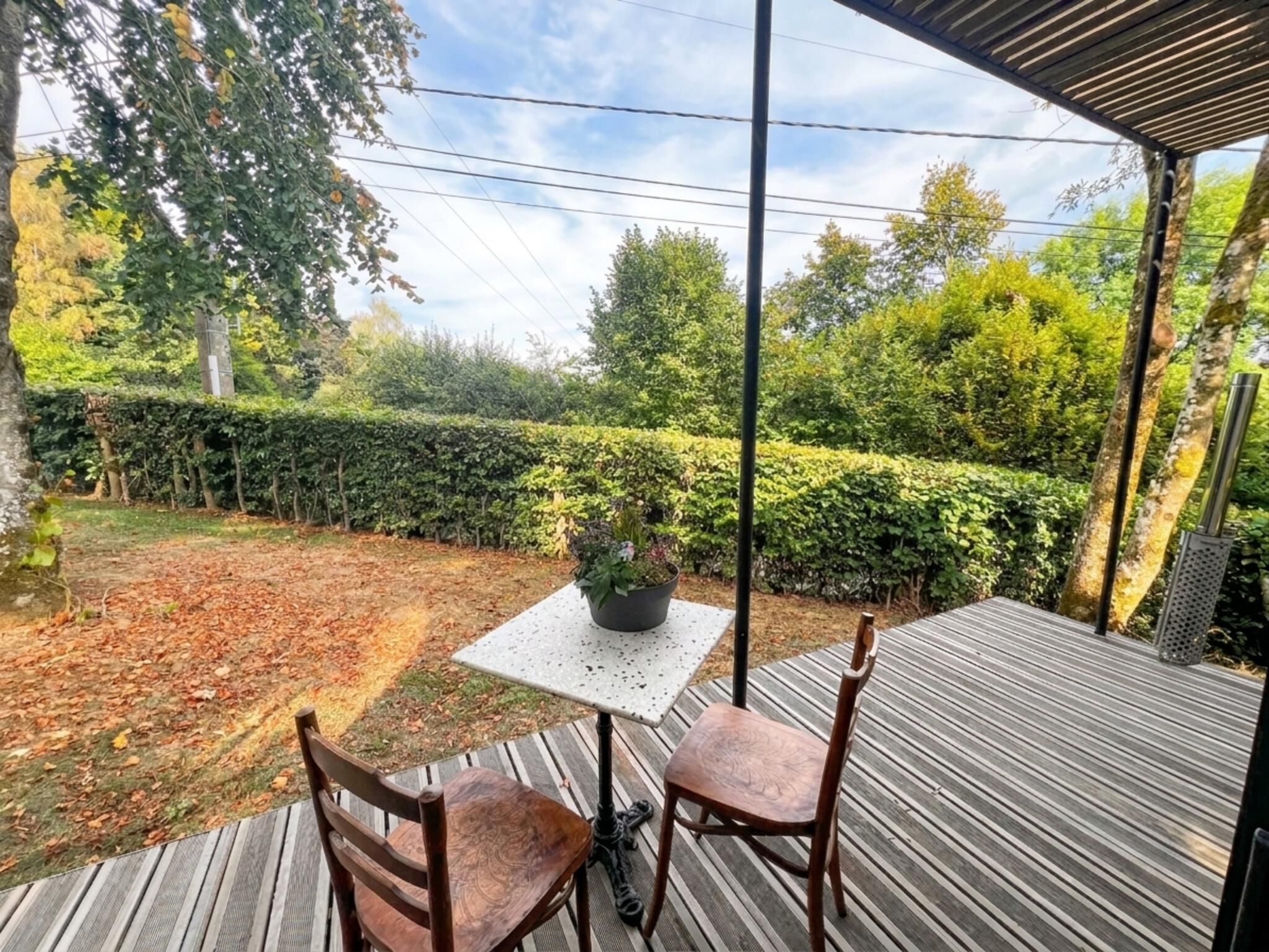 A covered outdoor seating area with a table and chairs, a potted plant, and a view of a landscaped garden.