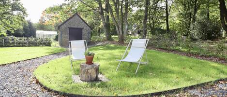 A garden with two white lawn chairs, a stone bench, and a small shed.