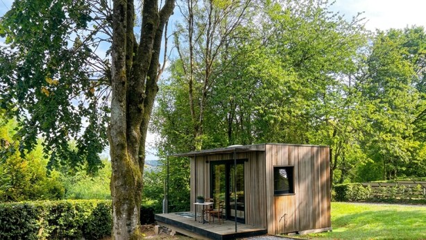 Una cabaña moderna de madera con puerta de vidrio, una pequeña terraza y un camino de grava que la conduce.