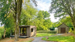 Un sendero de grava que lleva a una estructura de madera con puerta de cristal, rodeada de exuberante follaje y árboles.