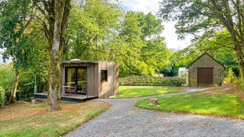 A gravel pathway leading to a wooden structure with a glass door, surrounded by lush greenery and trees.