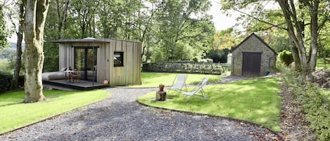 A gravel driveway leading to a wooden cabin with a deck and a stone building in the background.