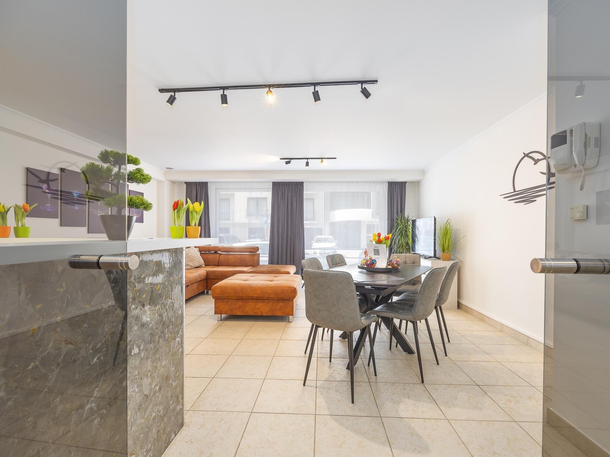 A modern living room with a sofa, dining table, and chairs. There is a kitchen area with a marble countertop and a built-in oven.
