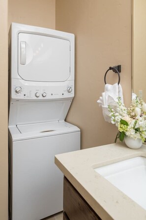 A compact, modern bathroom with a built-in washing machine, a towel rack, and a vase with flowers on the countertop. - Desert Bloom - Heated Pool & Spa, Sauna, Gym, Game Room (Sedona)