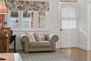 A room with a beige sofa, a patterned throw pillow, a striped rug, a white door, and a window with a white picket fence view. - Kalamunda Cottage (Kalamunda)