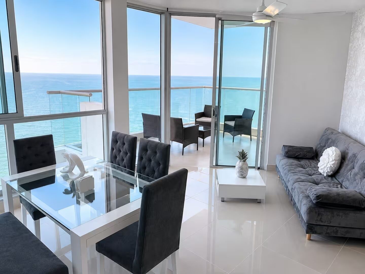 A modern living room with a glass table, chairs, a sofa, and a view of the sea.
