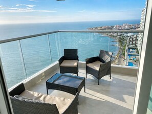 Ein Balkon mit Korbmöbeln und einem Glastisch, der Blick auf eine Küstenstadt und das Meer bietet.