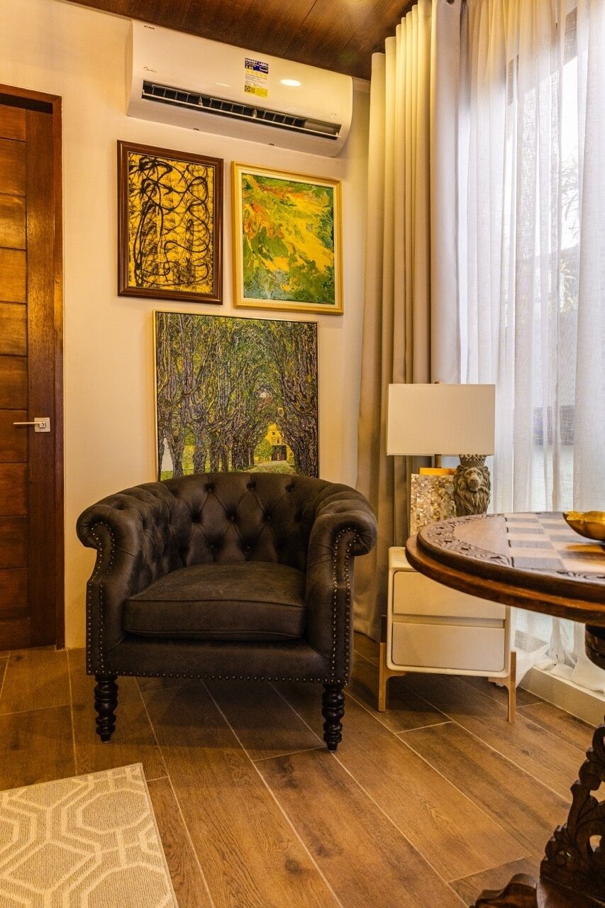 A room with a tufted armchair, a round table with a chessboard, and two framed artworks on the wall.