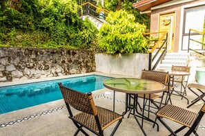 Un área de piscina con un muro de piedra, una mesa de vidrio, sillas de mimbre y una escalera que lleva a un edificio.