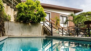 Ein Schwimmbecken mit Betonrand, einer Holzterrasse und einer Treppe zu einem Gebäude mit großen Fenstern.