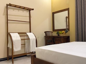 A hotel room with a bed, a wooden towel rack with towels, a wooden chair, a small wooden table with fruit, and a mirror on the wall.