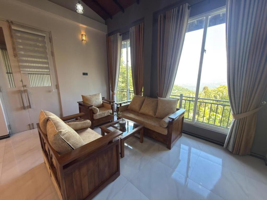 A spacious living room with a sofa set, a coffee table, and a view of greenery through large windows.