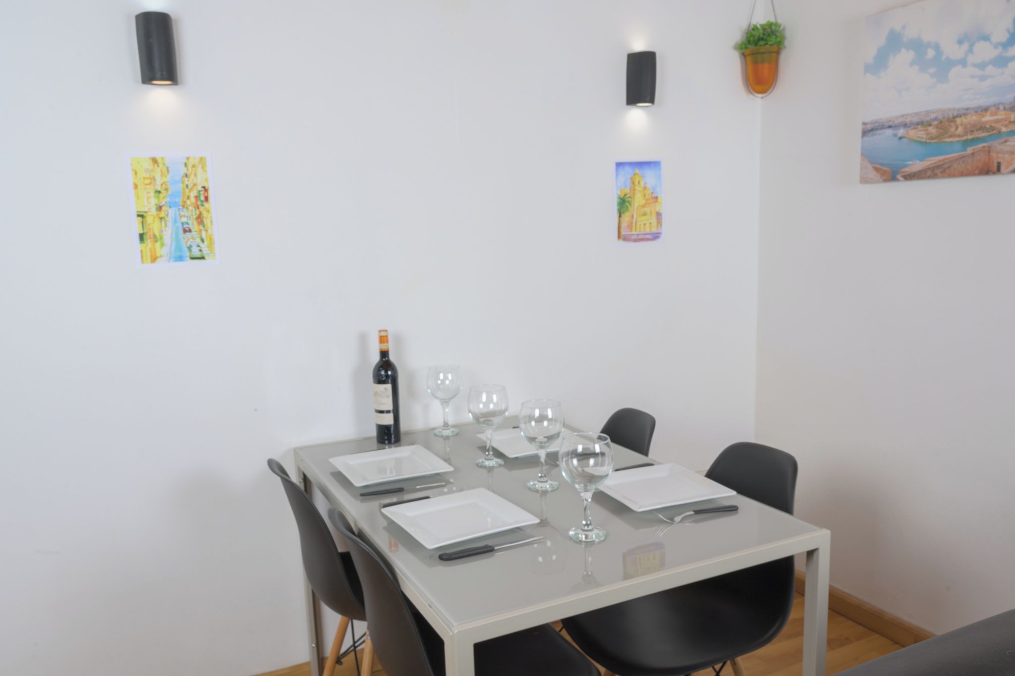 A dining area with a table set for six, a bottle of wine, and several wine glasses. There are black chairs around the table, and the room has white walls with a few artworks and a potted plant.