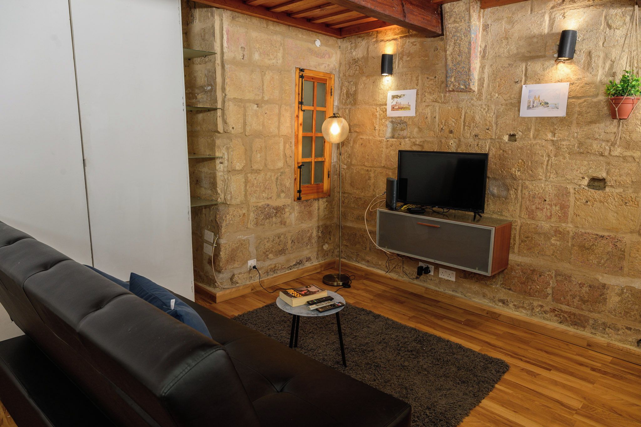 Una sala de estar acogedora con pared de piedra, techo de madera, sofá de cuero negro, televisor y una mesita con un libro y controles remotos.