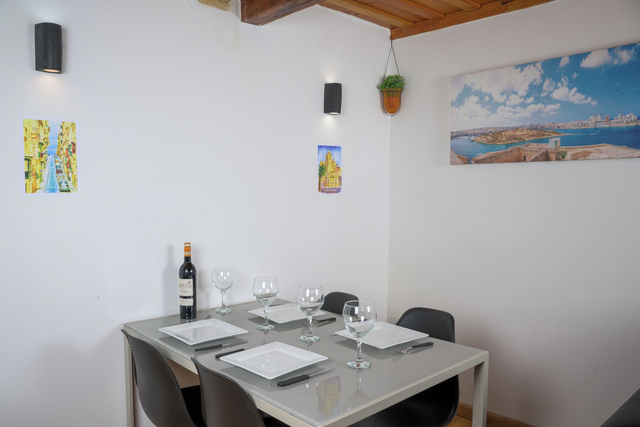 Un comedor con una mesa dispuesta para cuatro, una botella de vino y dos cuadros en la pared.