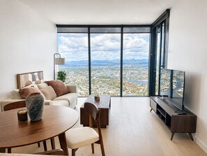 A modern living room with a large window, a sofa, a coffee table, and a TV. - Vista in the Clouds - Coastal City Escape (Broadbeach)
