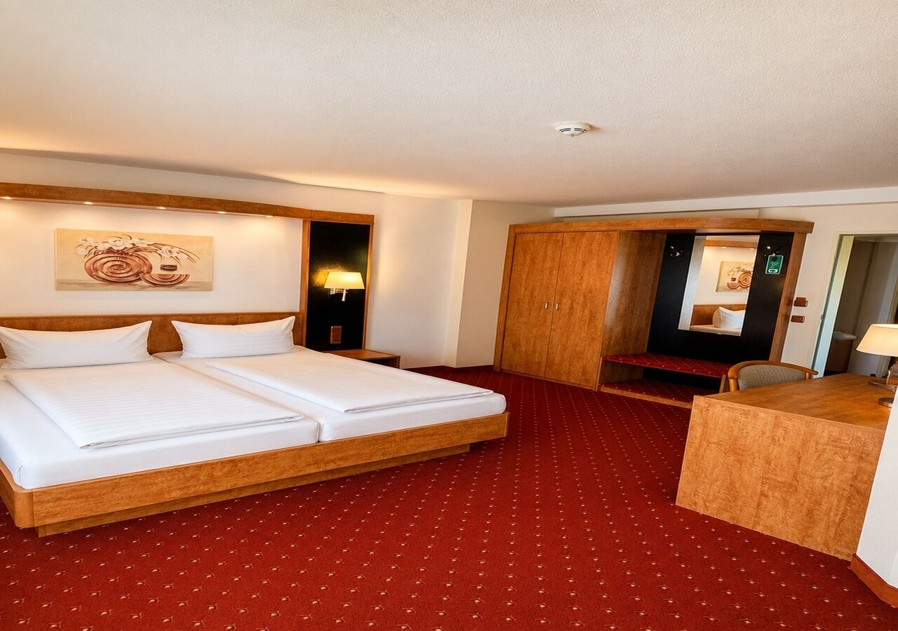 A hotel room with a double bed, a desk, a wardrobe, and a red carpeted floor.