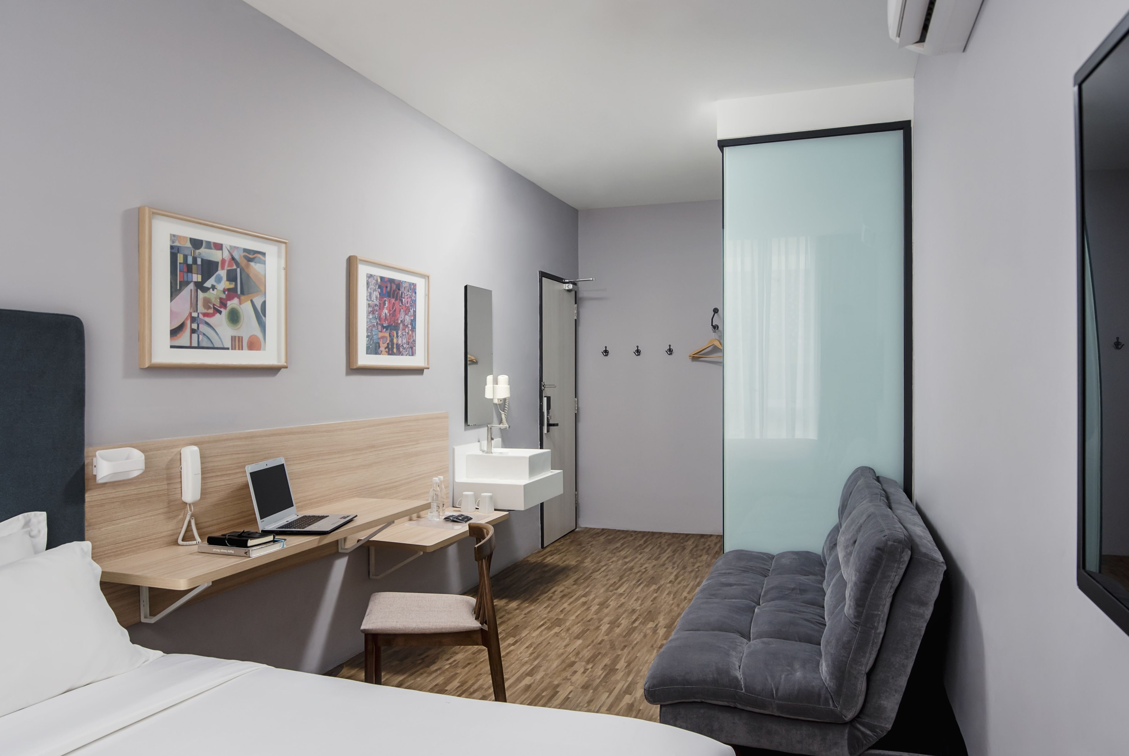 A hotel room with a bed, desk, chair, and a grey sofa. There are two framed artworks on the wall, a mirror, and a shower area.