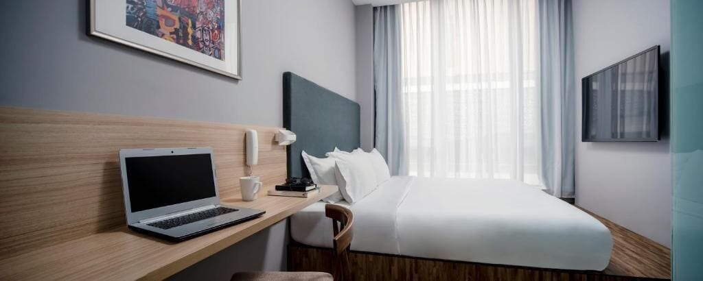 A hotel room with a bed, a desk with a laptop, a chair, a bedside table with a lamp, and a window with curtains.