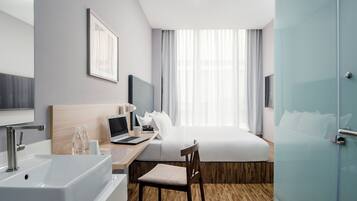 A hotel room with a bed, desk, laptop, and a bathroom with a sink and mirror.