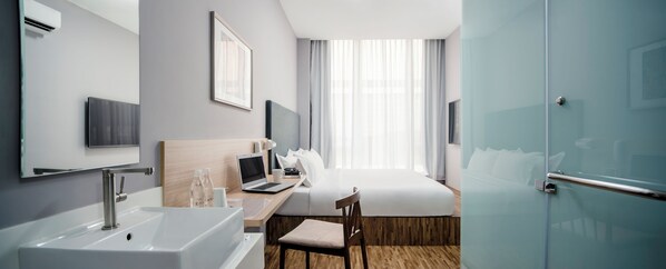 A hotel room with a bed, desk, laptop, and a bathroom with a sink and mirror.