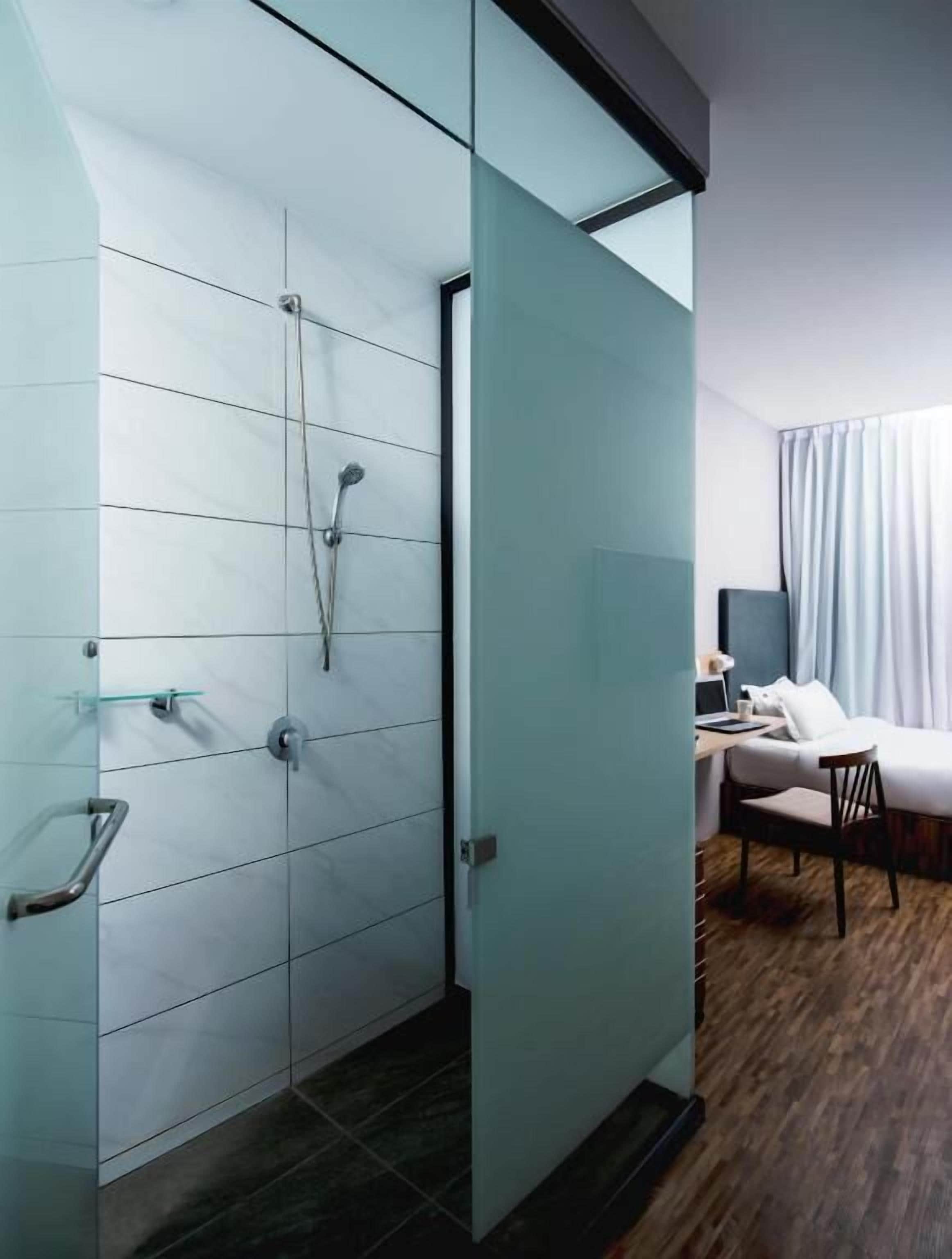 A modern bathroom with a glass shower enclosure, a wooden floor, and a bed with a white duvet in the adjacent room.