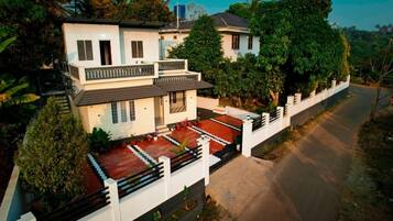 A two-story residential building with a balcony, surrounded by greenery and a paved driveway.
