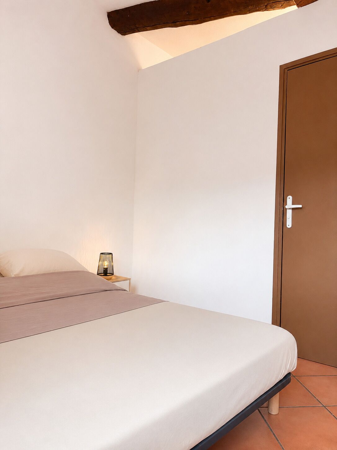 Een slaapkamer met een bed, nachtkastje, lamp en een deur.