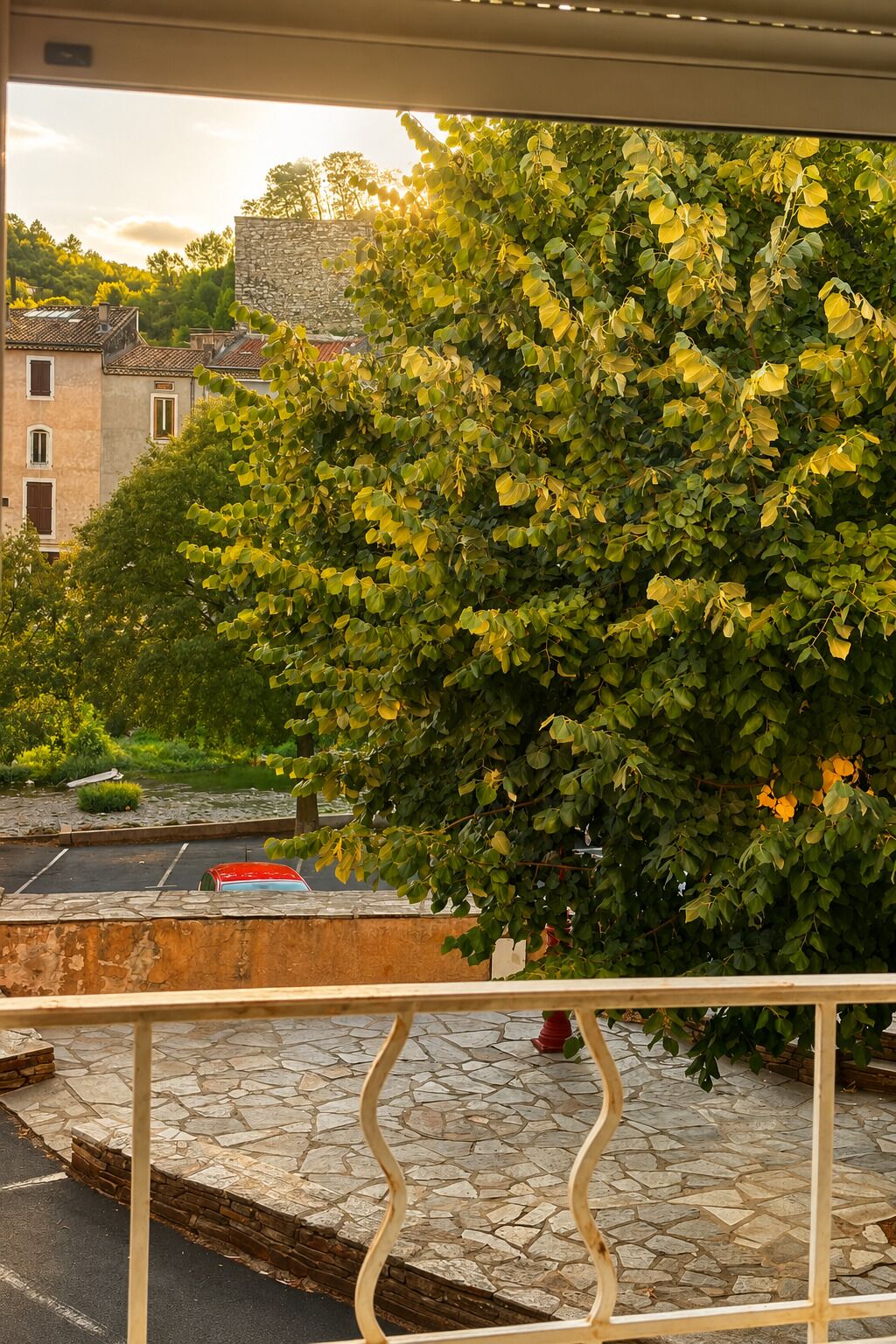 Un’area pavimentata in pietra con ringhiera metallica, un albero con foglie verdi e un edificio con tetto a tegole sullo sfondo.