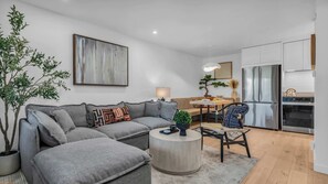 Ein modernes Wohnzimmer mit einem grauen Ecksofa, einem runden Couchtisch und einem Essbereich mit Holztisch und Stühlen.