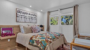 Ein Schlafzimmer mit Bett, Nachttisch, Fenster mit Vorhängen und einem Wandbild.