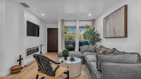Uma sala de estar moderna com um sofá cinzento, uma mesa de centro, uma TV fixada na parede e uma janela grande com vista para vegetação.