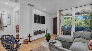 Ein modernes Wohnzimmer mit grauem Sofa, Flachbildfernseher, Kamin und einem großen Fenster mit Blick auf Grünflächen.