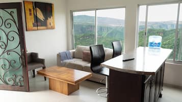 A modern living room with a sofa, a coffee table, and a dining area with a bar. There are large windows offering a view of a mountainous landscape.