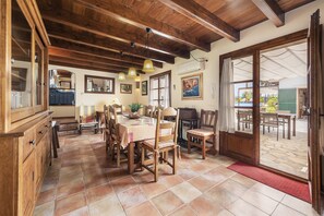 A dining area with wooden tables and chairs, a kitchen in the background, and a view of the outdoor patio through a glass door. - Grand Majorcan Retreat With Pool (Pollença)