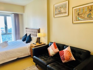 A hotel room with a bed, a black leather sofa, a nightstand with a lamp, and two framed pictures on the wall. - Studio Suite Condo at the Avida Towers Abreeza in vibrant Davao City (Davao City)