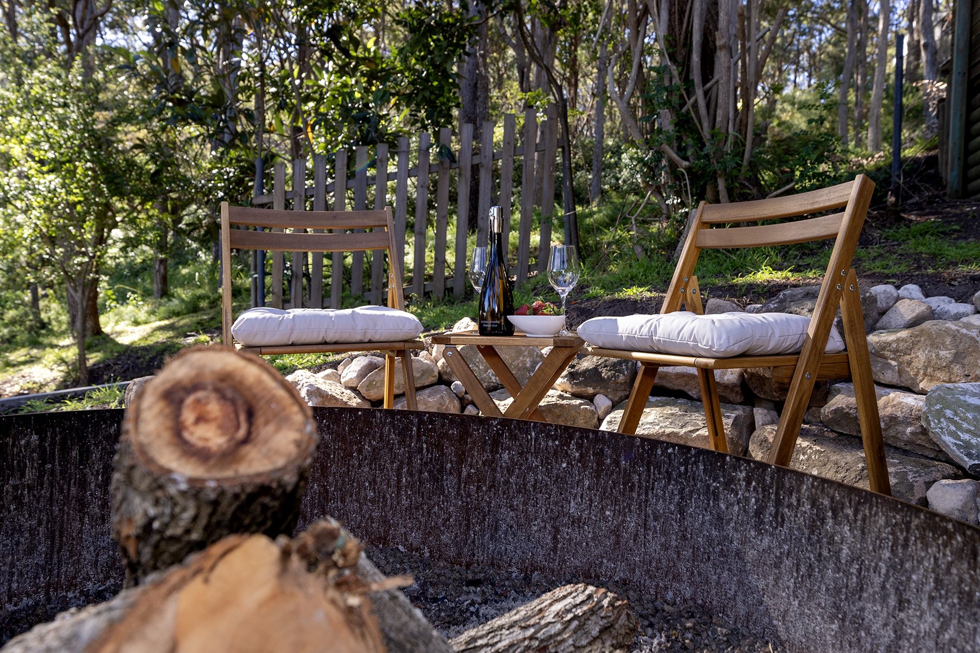 Two wooden chairs with cushions, a bottle of wine, and a bowl of fruit on a table in a natural setting with rocks and trees.