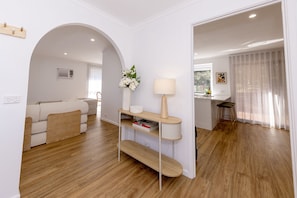 A modern living room with a sofa, a coffee table, and a lamp. There is a kitchen area visible through an archway. - Sunny 4BR Retreat: National Park, Surf & Beach (Forresters Beach)